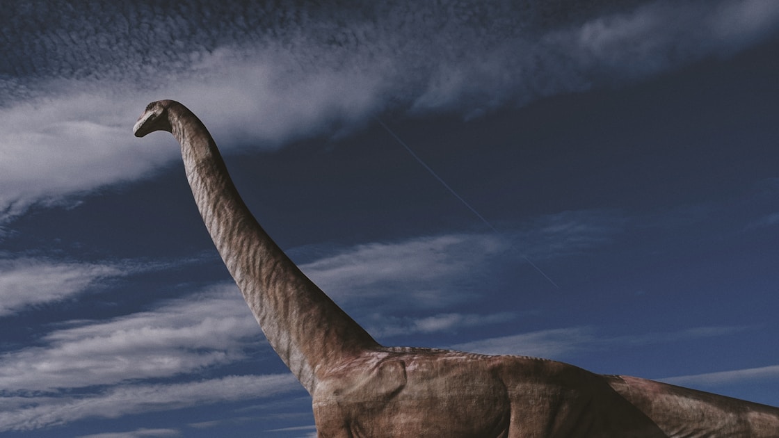 a large dinosaur walking across a field under a cloudy sky